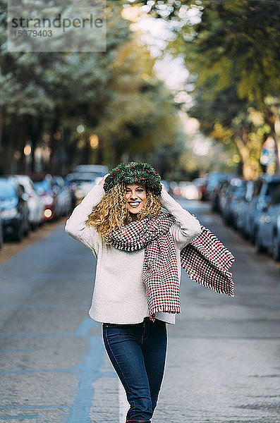 Porträt einer glücklichen jungen Frau  die mit einem Weihnachtskranz auf dem Kopf auf der Straße tanzt