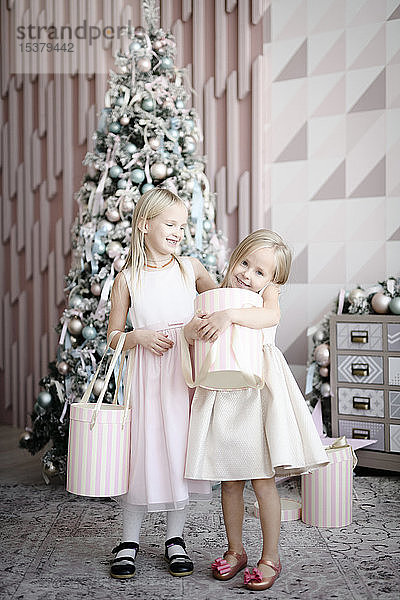 Porträt von zwei fröhlichen kleinen Mädchen  die vor einem beleuchteten Weihnachtsbaum mit Geschenkkartons stehen