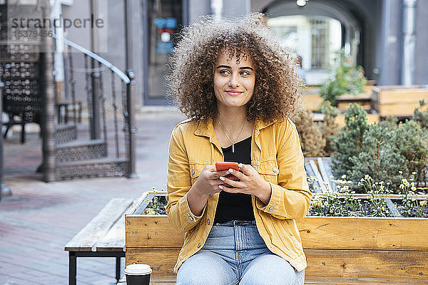 Porträt einer lächelnden Teenagerin  die mit Kaffee und Mobiltelefon auf einer Bank sitzt
