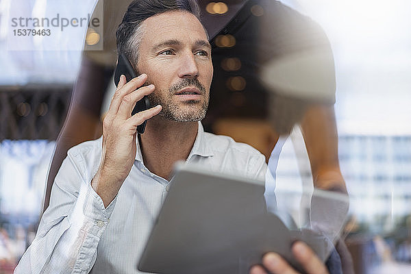 Porträt eines Geschäftsmannes mit Tablett am Telefon hinter einer Fensterscheibe in einem Cafe