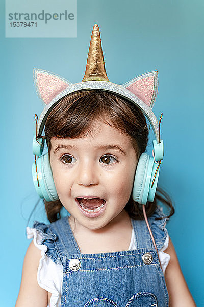 Porträt eines süßen kleinen Mädchens  das mit einhornförmigen Kopfhörern auf blauem Hintergrund Musik hört und singt
