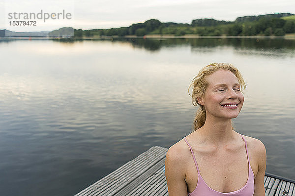 Lächelnde junge Frau mit geschlossenen Augen auf einem Steg an einem See