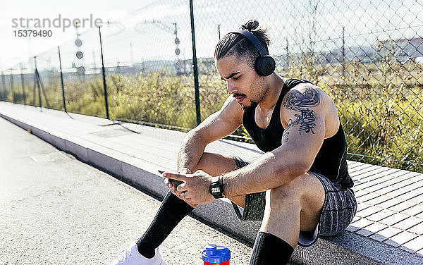 Sportlicher junger Mann mit Smartphone und Kopfhörern am Straßenrand sitzend