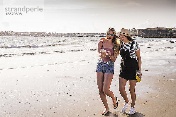 Zwei Freundinnen amüsieren sich  gehen am Strand spazieren  nehmen selbst ein Smartphone