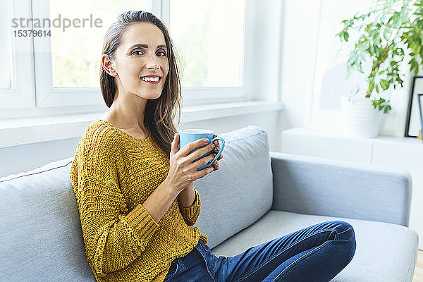 Porträt einer jungen Frau  die Kaffee trinkt  auf dem Sofa sitzt und in die Kamera lächelt