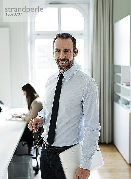 Porträt eines lächelnden Geschäftsmannes im Büro mit Mitarbeiter im Hintergrund