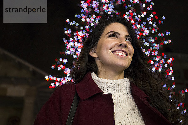 Porträt einer glücklichen jungen Frau vor beleuchtetem Weihnachtsbaum im Freien