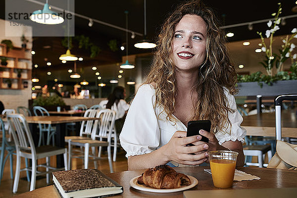 Lächelnde junge Frau mit Smartphone in einem Café beim Frühstück