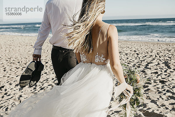 Rückansicht von Braut und Bräutigam beim Spaziergang am Strand