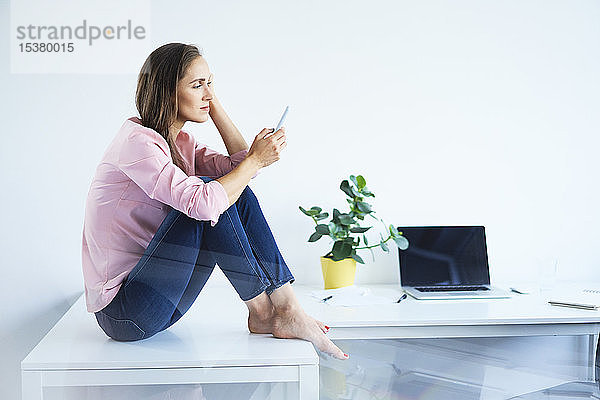 Junge Frau sitzt im Büro am Schreibtisch und benutzt ein Smartphone