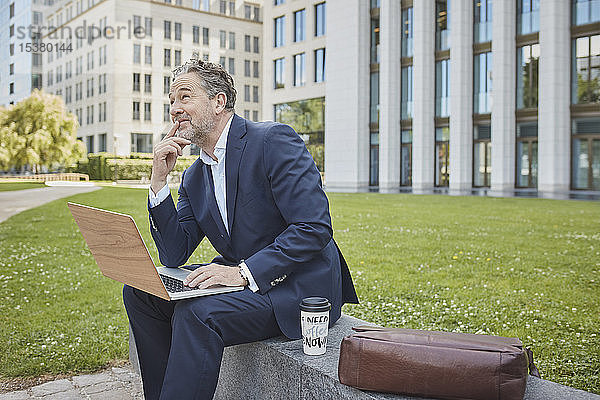 Reifer Geschäftsmann sitzt in der Stadt mit Laptop an einer Wand