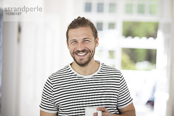 Porträt eines lächelnden jungen Mannes  der zu Hause eine Tasse Kaffee hält