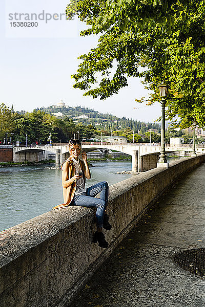 Frau mit Kopfhörern sitzt mit Smartphone am Flussufer