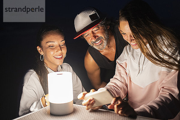 Zwei junge Frauen und ein reifer Mann mit Smartphone und LED-Licht auf nächtlichem Campingtisch