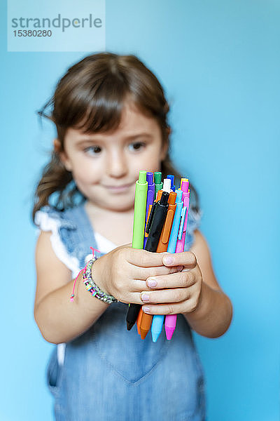 Porträt eines süßen kleinen Mädchens  das eine Handvoll farbiger Kugelschreiber auf blauem Hintergrund aufhebt