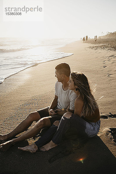 Glückliches junges Paar sitzt bei Sonnenuntergang am Strand