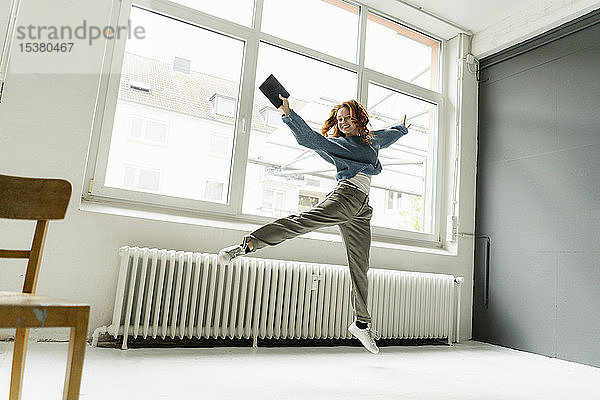 Porträt einer rothaarigen Frau mit digitalem Tablett  die auf einem Dachboden in die Luft springt