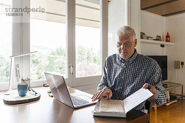 Älterer Mann sitzt zu Hause vor dem Laptop und blättert im Notizbuch um