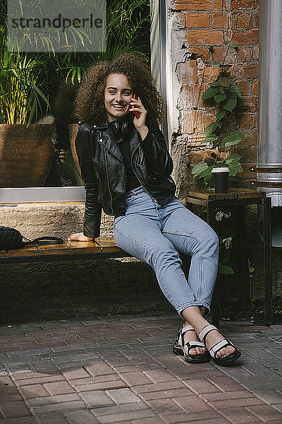 Porträt einer glücklichen Teenagerin am Telefon  die in einem Café im Freien mit Kaffee zum Mitnehmen sitzt