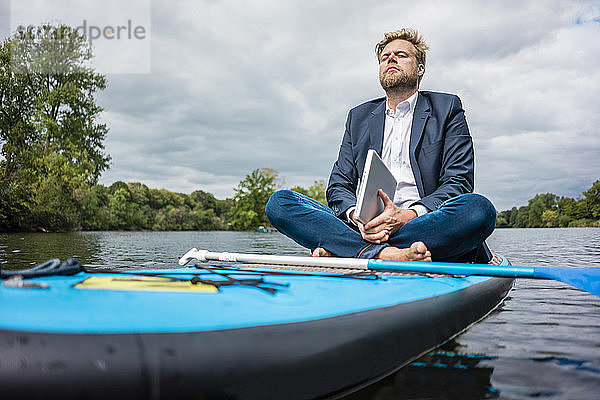 Geschäftsmann mit Laptop sitzt auf einem SUP-Brett auf einem See