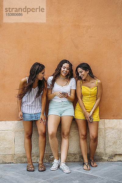 Drei glückliche Freundinnen stehen an einer Wand und teilen sich ein Smartphone