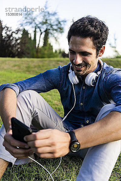 Lächelnder Mann sitzt auf einer Wiese und benutzt sein Smartphone