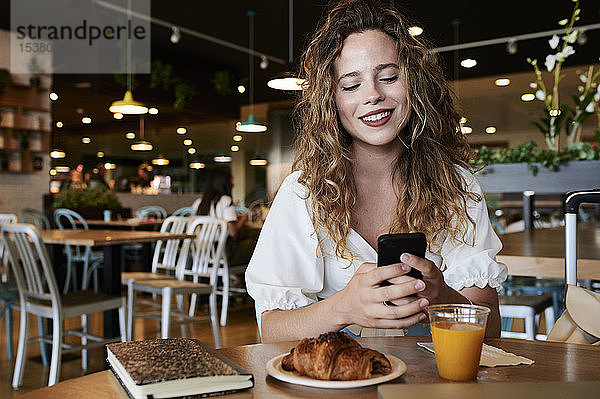 Lächelnde junge Frau benutzt Smartphone in einem Café während des Frühstücks