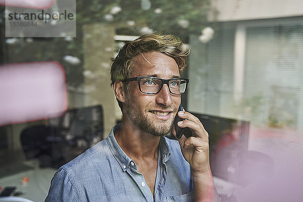 Lässiger junger Geschäftsmann am Telefon hinter einer Fensterscheibe im Büro