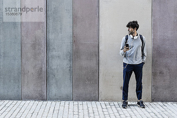 Mann mit Handy und Kopfhörern vor einer Mauer stehend