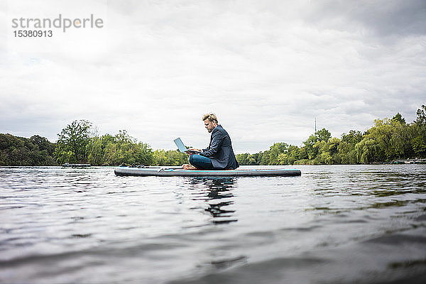 Geschäftsmann sitzt mit Laptop auf einem SUP-Brett auf einem See