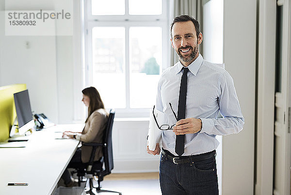 Porträt eines lächelnden Geschäftsmannes im Büro mit Mitarbeiter im Hintergrund