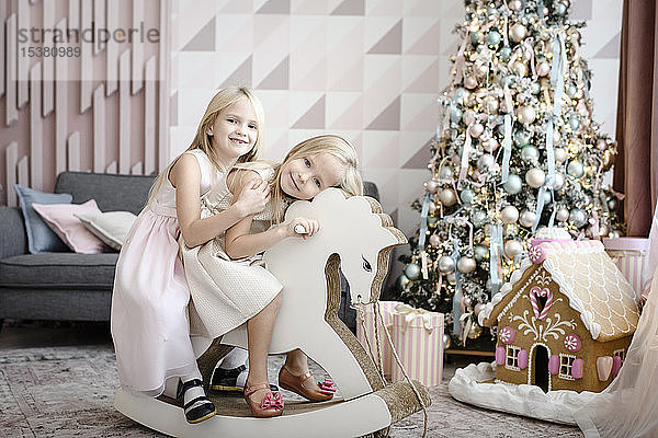 Porträt von zwei glücklichen kleinen Mädchen zusammen auf einem Schaukelpferd zur Weihnachtszeit