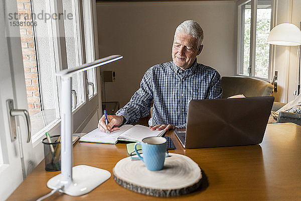 Älterer Mann sitzt zu Hause vor dem Laptop und macht Notizen im Notizbuch