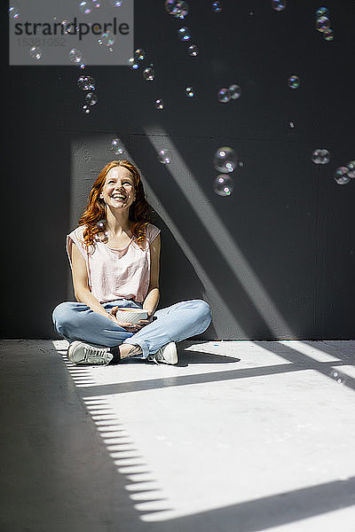 Porträt einer glücklichen Frau  die mit einer Kaffeeschüssel auf dem Boden sitzt und Seifenblasen betrachtet