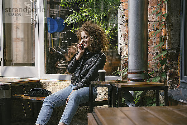 Glückliches Teenagermädchen am Telefon sitzt in einem Café im Freien mit Kaffee zum Mitnehmen