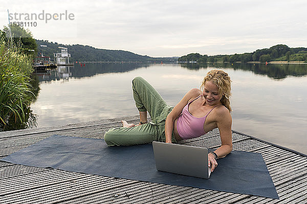 Lächelnde junge Frau mit Laptop auf einem Steg an einem See