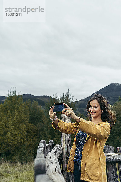 Junge Frau in gelbem Mantel beim Fotografieren mit ihrem Smartphone