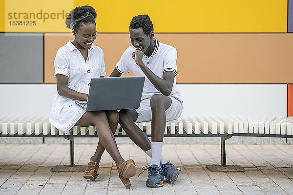 Lächelndes junges Paar sitzt auf der Bank und schaut auf den Laptop