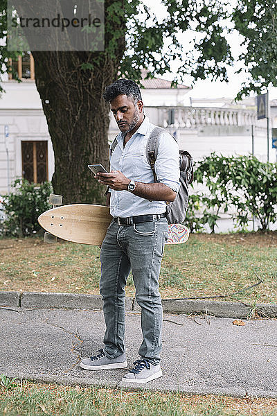 Gelegenheits-Geschäftsmann mit Skateboard mit Smartphone in der Stadt