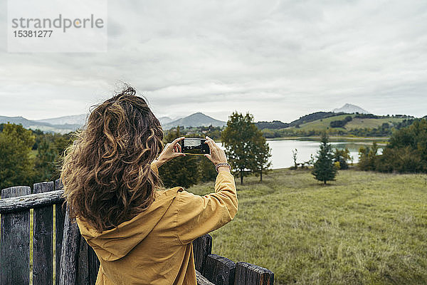 Junge Frau in gelbem Mantel beim Fotografieren mit ihrem Smartphone