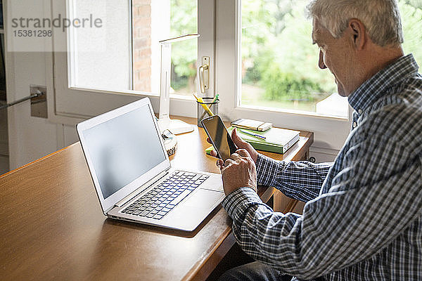Älterer Mann sitzt zu Hause vor dem Laptop und benutzt ein Smartphone