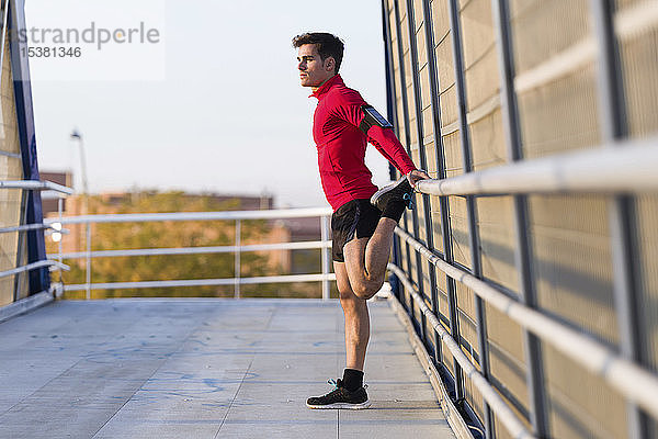 Jogger mit Smartphone in der Armtasche  streckt sein Bein auf einem Brückengeländer