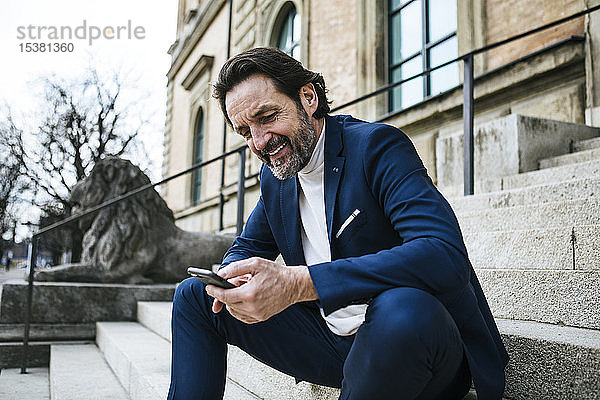 Porträt eines lächelnden reifen Geschäftsmannes  der auf einer Treppe sitzt und auf sein Handy schaut