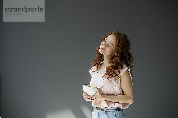 Porträt einer glücklichen rothaarigen Frau mit Kaffeeschüssel vor grauem Hintergrund