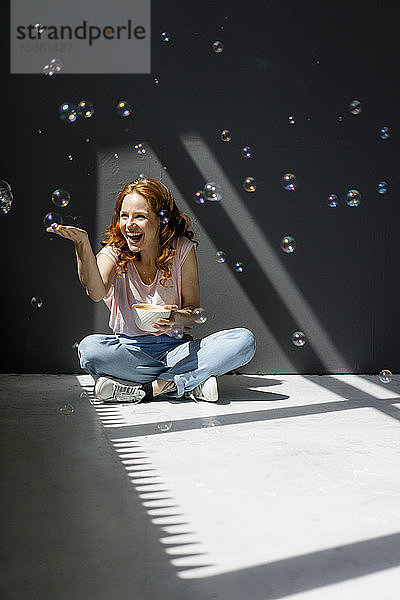 Porträt einer glücklichen Frau  die mit einer Kaffeeschüssel auf dem Boden sitzt und Seifenblasen berührt