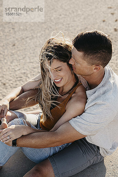 Glückliches  anhängliches junges Paar sitzt am Strand