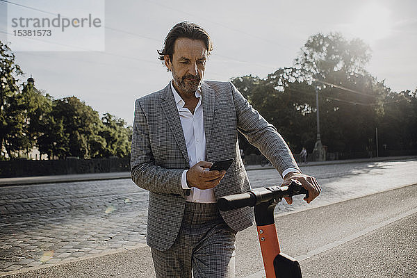 Geschäftsmann schaut auf Smartphone mit E-Scooter in der Stadt