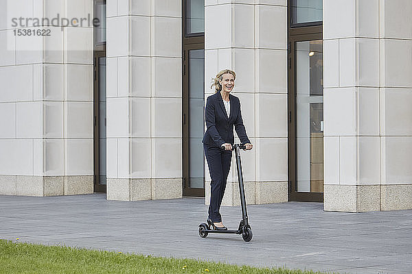 Geschäftsfrau auf E-Scooter fährt an Bürogebäude in der Stadt vorbei