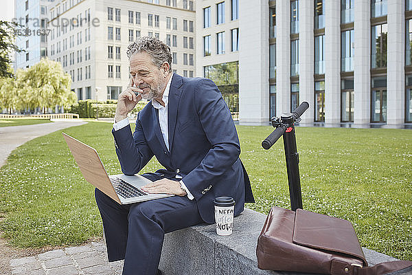 Reifer Geschäftsmann sitzt in der Stadt mit Laptop an einer Wand