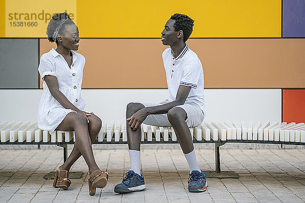 Lächelndes junges Paar sitzt auf einer Bank und sieht sich an